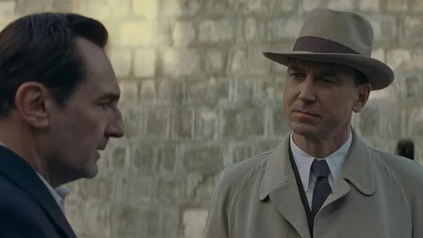 Gilles Lellouche, portant un fedora et un trench-coat, en conversation avec un autre homme en costume sombre devant un mur en pierre, dans une scène du film "Moulin".