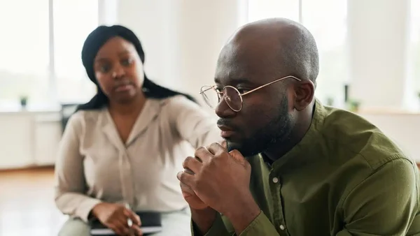 Un homme noir en chemise verte et une femme noire en chemise beige assise, dans une pièce lumineuse, échangeant un regard chargé de tension, illustrant le silence entre partenaires