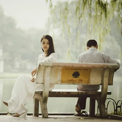 Une jeune femme en robe blanche assise sur un banc de parc, regardant la caméra, tandis qu'un homme assis à côté de lui tourne le dos, face à un lac brumeux, illustrant l'éloignement émotionnel