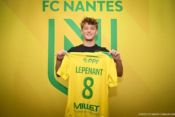 Johann Lepenant, joueur prêté par l'OL à Nantes, sourit en montrant son maillot jaune avec le nom 'LEPENANT' et le numéro 8 devant le mur jaune et le logo de FC Nantes
