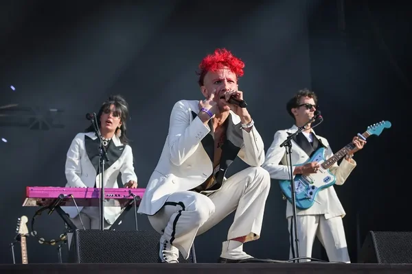 La Femme interprète "Sacatela" sur scène : un chanteur aux cheveux roux, vêtu de blanc, chante au micro devant un claviériste et un guitariste également en blanc.