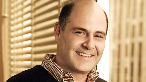 Portrait en gros plan de Matthew Weiner, souriant, portant une chemise à carreaux et un pull sombre, devant des stores à lamelles.