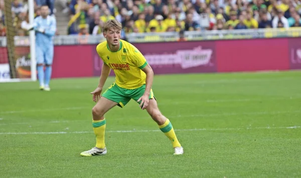 Johann Lepenant, joueur de football en maillot jaune et vert, en position défensive sur un terrain en herbe pendant un match de Ligue 1 avec des spectateurs en arrière-plan