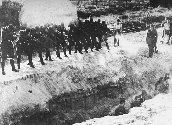 Photographie en noir et blanc montrant une ligne de soldats pointant des fusils vers un groupe de personnes debout dans une tranchée, illustrant la Shoah par balles commise en Lituanie.