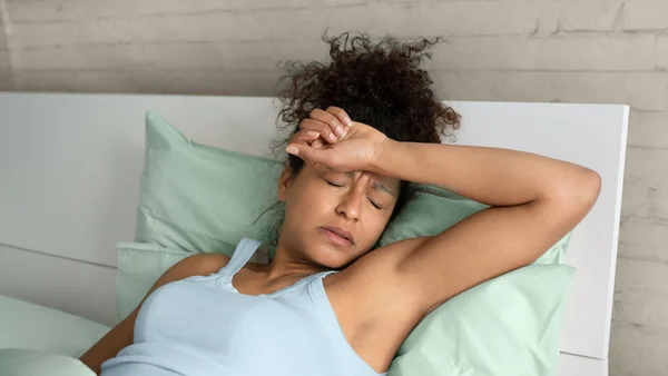 Une femme aux cheveux bouclés allongée dans son lit, la main posée sur le front, l'air souffrant, pour illustrer les sueurs nocturnes et les difficultés de sommeil liées à la ménopause.