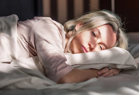 Une femme blonde allongée sur le côté dans son lit, la tête reposant sur un oreiller, les yeux ouverts et l'air pensif, illustrant les troubles du sommeil et les bouffées de chaleur liés à la ménopause.
