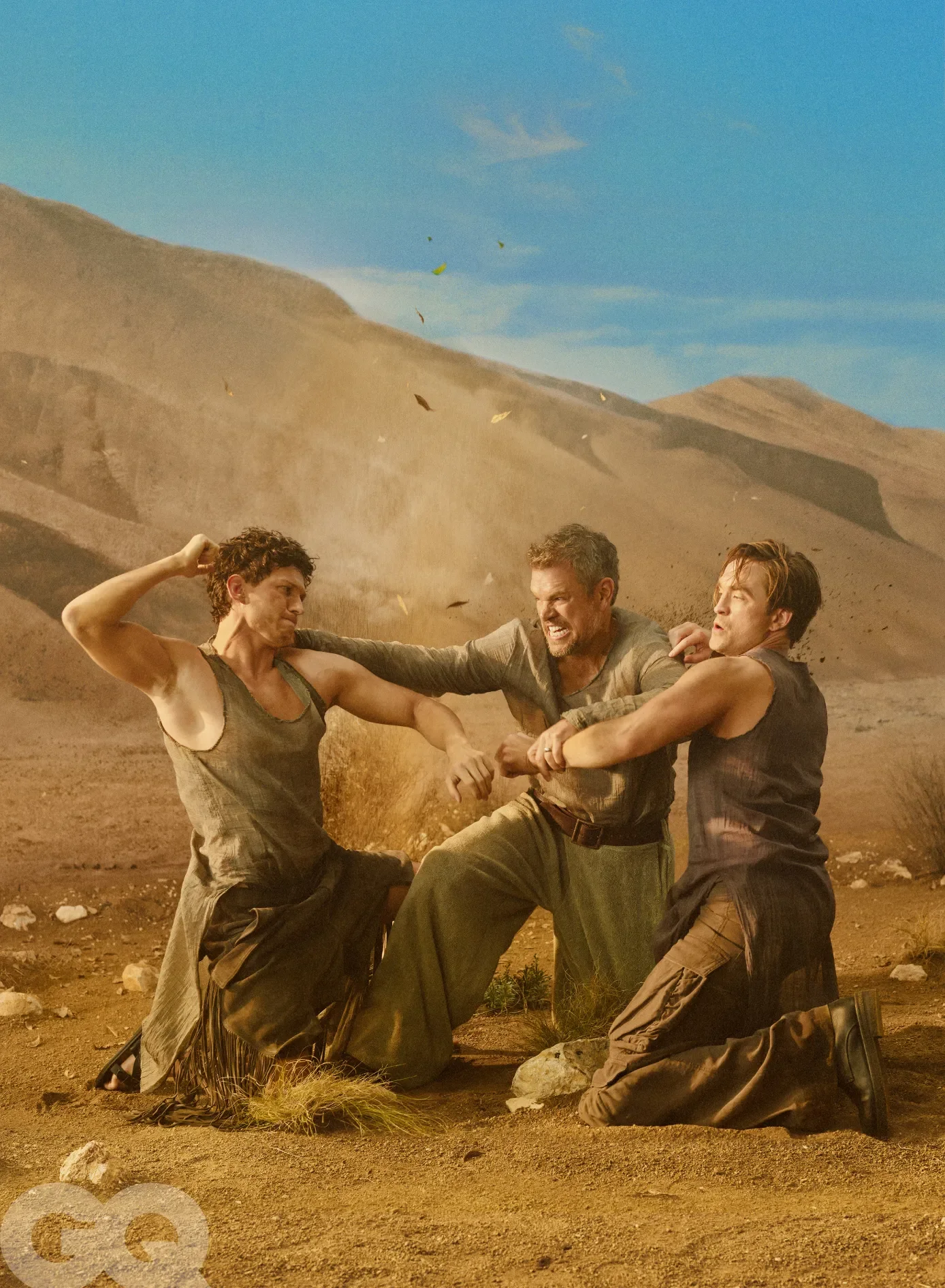 Une photographie de style éditorial montrant les acteurs Tom Holland et Robert Pattinson, ainsi que le réalisateur Christopher Nolan, dans un décor désertique. Les trois hommes sont agenouillés sur le sol, vêtus de costumes rappelant l'Antiquité grecque. L'image capture une scène d'action dynamique où Holland et Pattinson semblent se battre ou interagir physiquement sous la direction de Nolan, avec du sable et des débris volant en l'air. Le logo du magazine GQ est visible en bas à gauche.