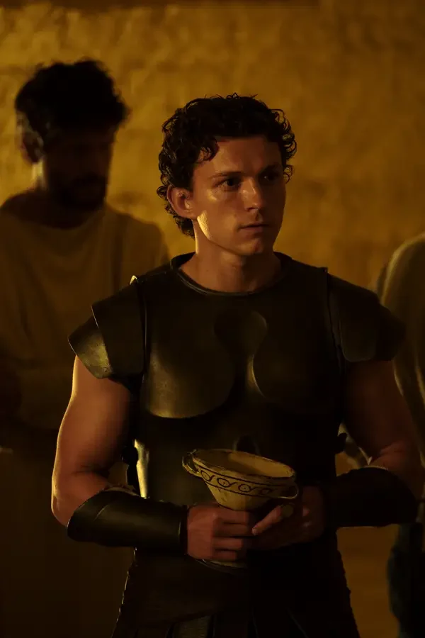 L'image montre l'acteur Tom Holland dans un costume d'époque, ressemblant à une armure grecque antique. Il tient un bol en céramique et a une expression sérieuse. L'éclairage est chaud et la scène semble se dérouler dans un intérieur ancien. Un autre personnage est visible mais flou en arrière-plan.
